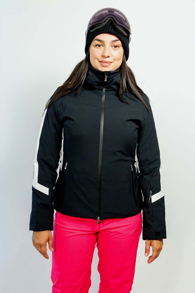 AthenaWomensInsulatedJacket-A woman modeling a black ski jacket with white sleeve stripes, bright pink ski pants, a black beanie, and ski goggles, set against a plain light gray studio background.