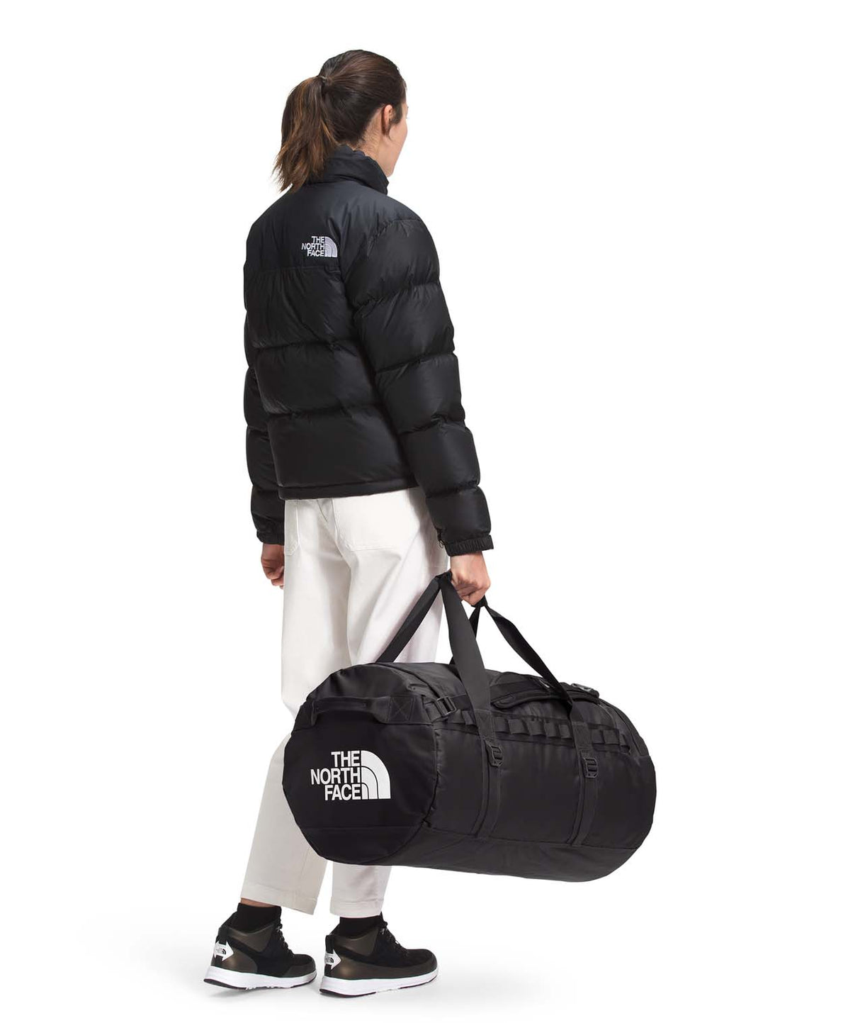 BaseCampDuffel_M-Person wearing a black jacket and white pants, carrying a black duffel bag with The North Face logo on a white background.