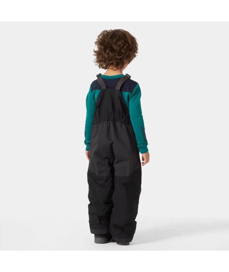 KidsRider2InsulatedBib- A young child wearing black Helly Hansen snow bib overalls over a teal long-sleeve shirt, standing against a light gray studio background.