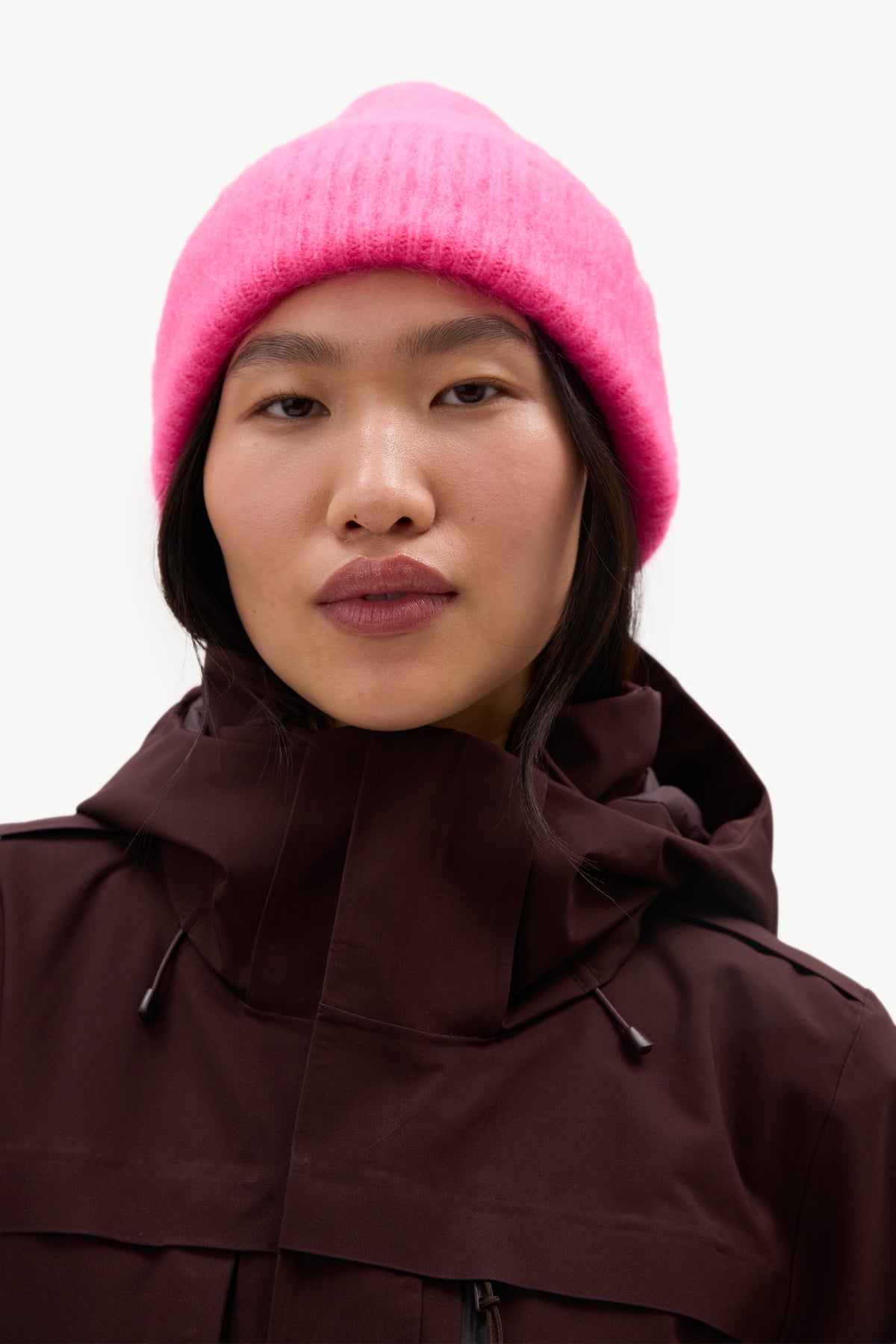 MOHAIRBEANIE- A close-up portrait of a model wearing a bright pink knit beanie and a dark brown hooded jacket against a plain light background.
