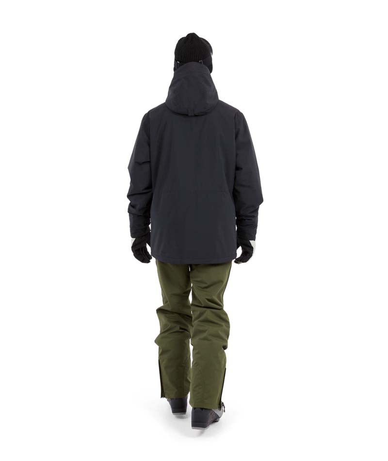 NateJacket-A man wearing a black XTM snowboard jacket, black beanie with green-tinted goggles, black gloves, and olive pants, standing against a solid white background.