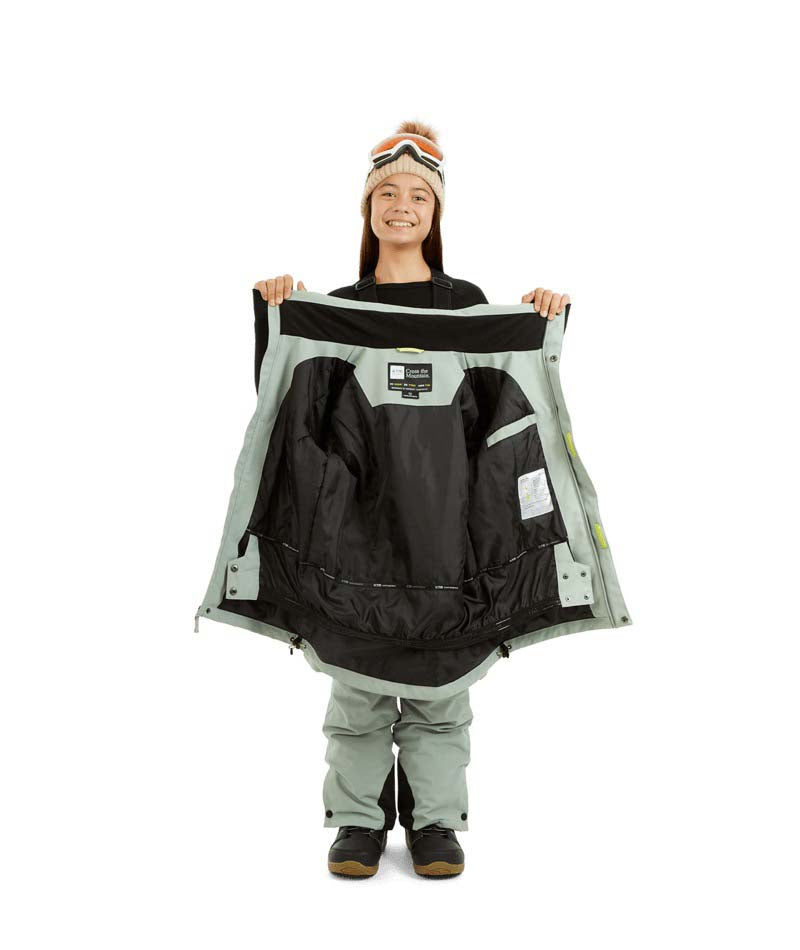 RumbleJacket- A smiling girl wearing a light sage green snowboard jacket, matching snow pants, black gloves, and a knit beanie with ski goggles, standing against a plain white background.