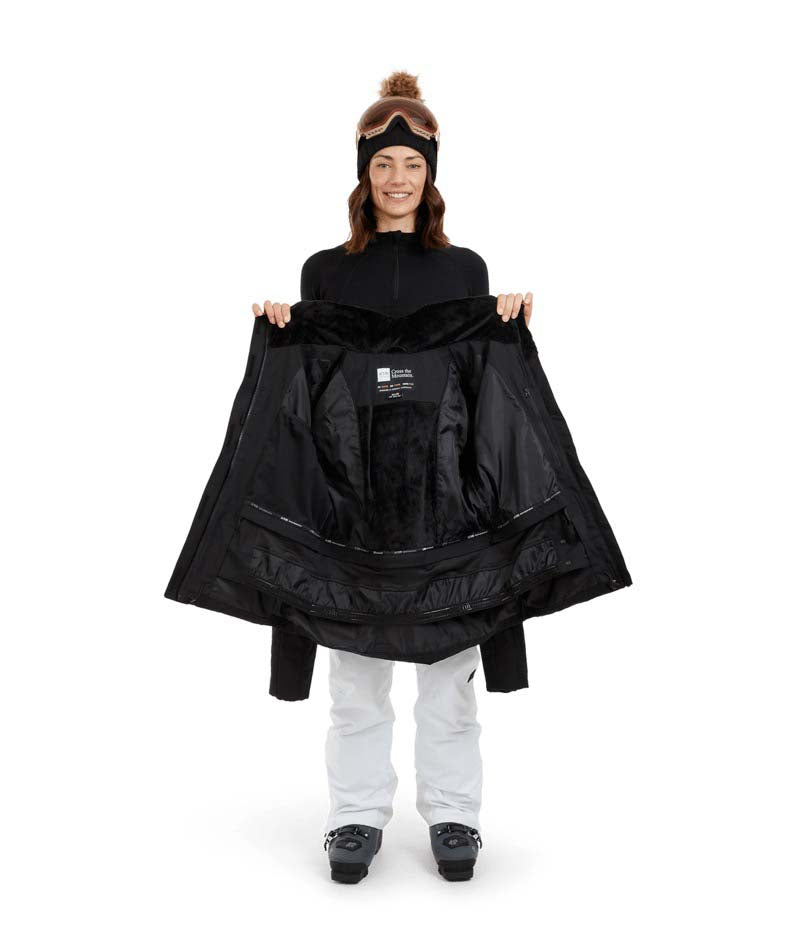 SolaceJacket-A woman wearing a black ski jacket with a faux-fur trimmed hood, black gloves, a black beanie, and ski goggles, posed against a solid white background.