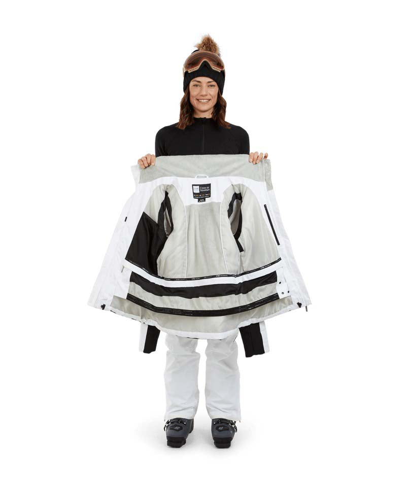 SolaceJacket-A woman wearing a white and black ski jacket with a black fur-trimmed hood, black gloves, a black beanie, and ski goggles, posed against a solid white background.
