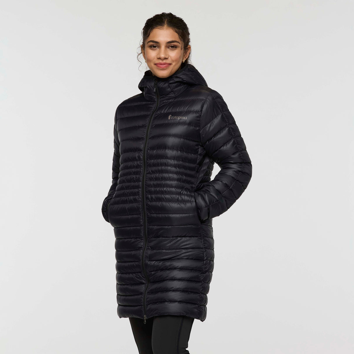 w-fuego-down-parka-A woman modeling a long, black quilted puffer jacket with a hood and a small embroidered 'Cotopaxi' logo on the left chest, set against a plain light gray studio background.