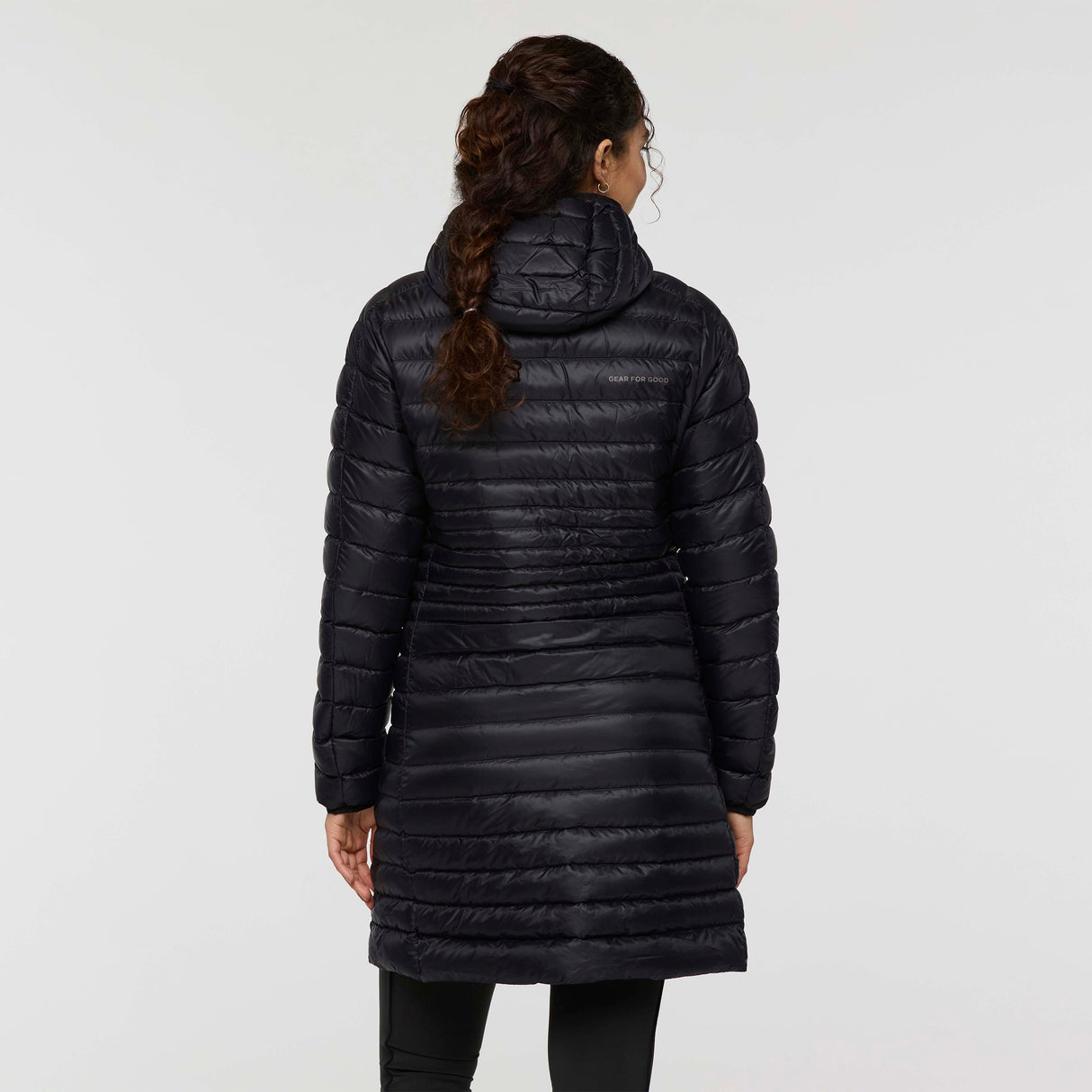w-fuego-down-parka-A woman modeling a long, black quilted puffer jacket with a hood and a small embroidered 'Cotopaxi' logo on the left chest, set against a plain light gray studio background.