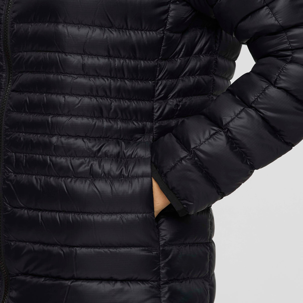 w-fuego-down-parka-A woman modeling a long, black quilted puffer jacket with a hood and a small embroidered 'Cotopaxi' logo on the left chest, set against a plain light gray studio background.