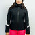 AthenaWomensInsulatedJacket-A woman modeling a black ski jacket with white sleeve stripes, bright pink ski pants, a black beanie, and ski goggles, set against a plain light gray studio background.