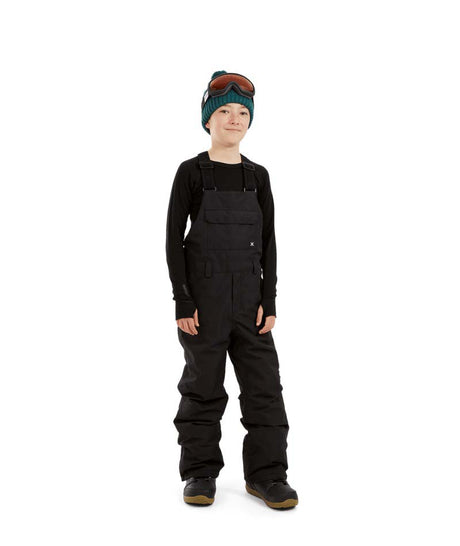 BaileyPant-A child model wearing black snow bib overalls, a black long-sleeve shirt, a dark green beanie with ski goggles, and black snow boots, standing against a solid white background.