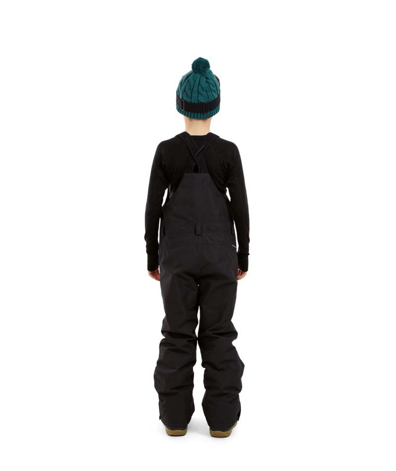 BaileyPant-A child model wearing black snow bib overalls, a black long-sleeve shirt, a dark green beanie with ski goggles, and black snow boots, standing against a solid white background.