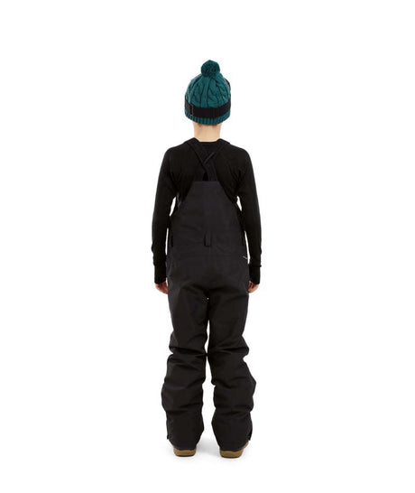 BaileyPant-A child model wearing black snow bib overalls, a black long-sleeve shirt, a dark green beanie with ski goggles, and black snow boots, standing against a solid white background.