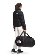 BaseCampDuffel_M-Person wearing a black jacket and white pants, carrying a black duffel bag with The North Face logo on a white background.
