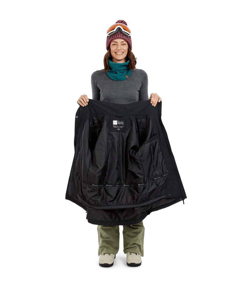EmeryJacket-A woman wearing a black hooded snowboard jacket, gray gloves, olive green pants, and a maroon knit beanie with ski goggles, standing against a plain white background.