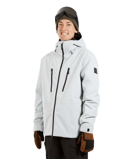 ExposureJacket-A man wearing a light gray ski jacket with black zippers, a dark beanie with goggles, and brown gloves, standing against a plain white background.