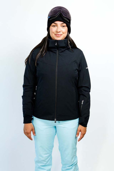 GaiaWomensInsulatedJacket-A woman modeling a black ski jacket with a high collar and front zipper, paired with light blue ski pants and a dark beanie with goggles, against a plain white studio background.
