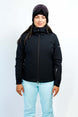 GaiaWomensInsulatedJacket-A woman modeling a black ski jacket with a high collar and front zipper, paired with light blue ski pants and a dark beanie with goggles, against a plain white studio background.