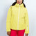 GaiaWomensInsulatedJacket-A woman modeling a bright yellow ski jacket with a white zipper, paired with red ski pants, blue goggles, and a black beanie against a light gray studio background.