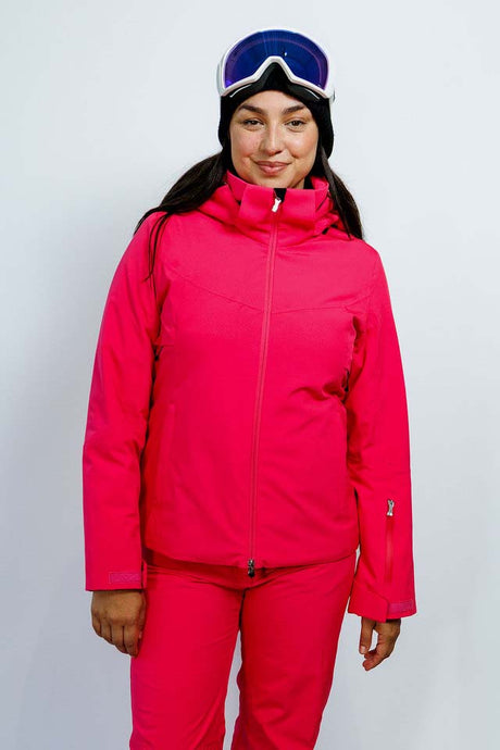 GaiaWomensInsulatedJacket-A woman modeling a bright pink ski jacket and matching ski pants, wearing a black beanie, dark goggles, and a black headband against a plain light gray studio background.