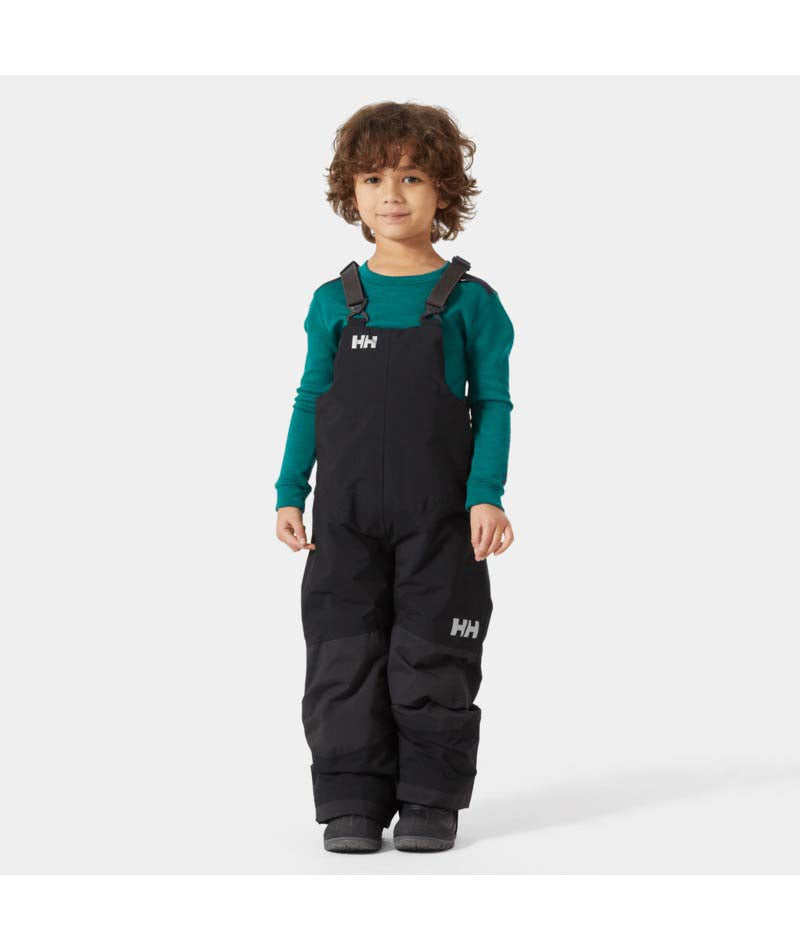 KidsRider2InsulatedBib- A young child wearing black Helly Hansen snow bib overalls over a teal long-sleeve shirt, standing against a light gray studio background.