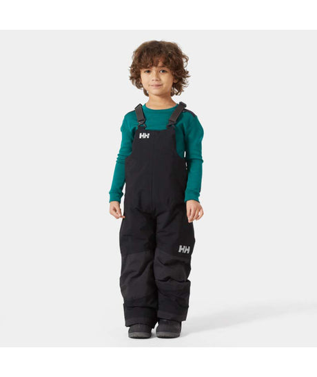 KidsRider2InsulatedBib- A young child wearing black Helly Hansen snow bib overalls over a teal long-sleeve shirt, standing against a light gray studio background.