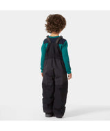 KidsRider2InsulatedBib- A young child wearing black Helly Hansen snow bib overalls over a teal long-sleeve shirt, standing against a light gray studio background.