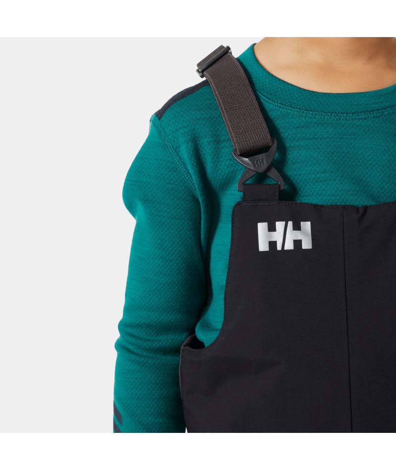 KidsRider2InsulatedBib- A young child wearing black Helly Hansen snow bib overalls over a teal long-sleeve shirt, standing against a light gray studio background.