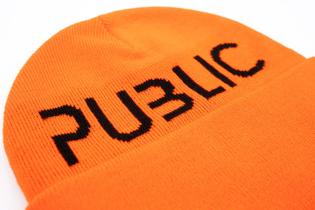LogoCuffBeanie-A close-up of a bright orange knit beanie with the word 'PUBLIC' embroidered in black across the front fold, shown on a plain white background.