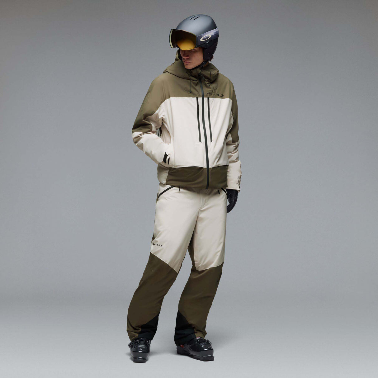MCanopyProInsulatedJacket-A color-blocked hooded jacket with an olive green upper section, hood, and lower front panel, paired with off-white color-blocked sleeves and a lower front panel. It features a black front zipper, black drawstrings, and a small Oakley logo on the chest.
