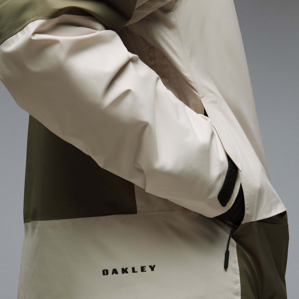 MCanopyProInsulatedJacket-A color-blocked hooded jacket with an olive green upper section, hood, and lower front panel, paired with off-white color-blocked sleeves and a lower front panel. It features a black front zipper, black drawstrings, and a small Oakley logo on the chest.