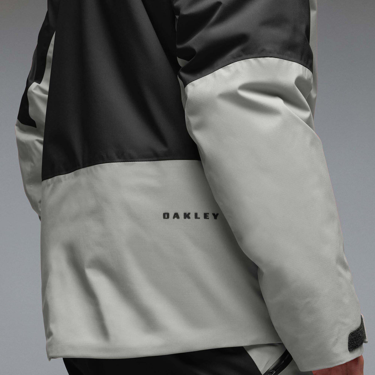 MCanopyProInsulatedJacket-A colorblock hooded jacket with a black upper section and sleeves, a light gray midsection, and a black lower section, featuring a full-zip front and a small white Oakley logo on the chest.
