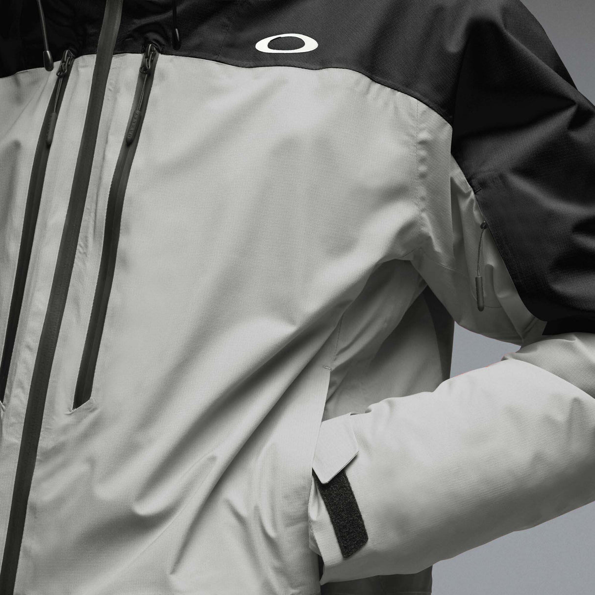 MCanopyProInsulatedJacket-A colorblock hooded jacket with a black upper section and sleeves, a light gray midsection, and a black lower section, featuring a full-zip front and a small white Oakley logo on the chest.