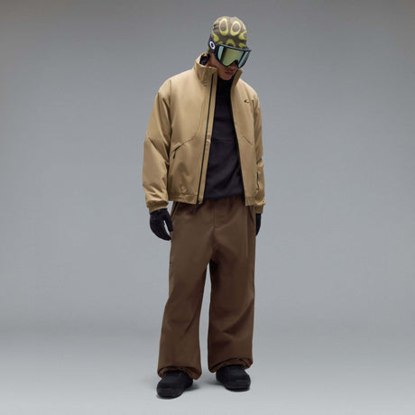 MChannelJacket-A beige lightweight zip-up jacket with a black front zipper, black side zip pockets, and a small black Oakley logo on the left chest, displayed against a neutral gray gradient background.