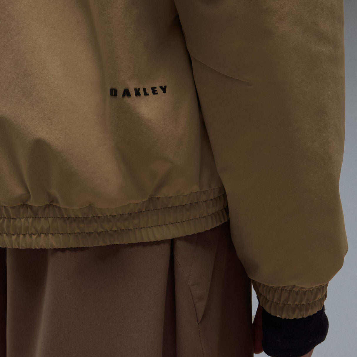 MChannelJacket-A beige lightweight zip-up jacket with a black front zipper, black side zip pockets, and a small black Oakley logo on the left chest, displayed against a neutral gray gradient background.