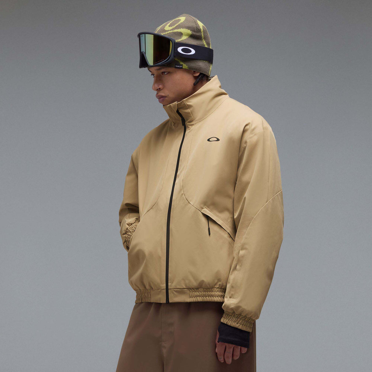 MChannelJacket-A beige lightweight zip-up jacket with a black front zipper, black side zip pockets, and a small black Oakley logo on the left chest, displayed against a neutral gray gradient background.