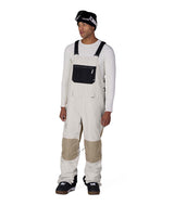 MHotLapInsulatedBib- A man modeling off-white snow bib overalls with beige knee patches, worn over a white long-sleeve shirt and paired with black snow boots and goggles, on a solid white background.