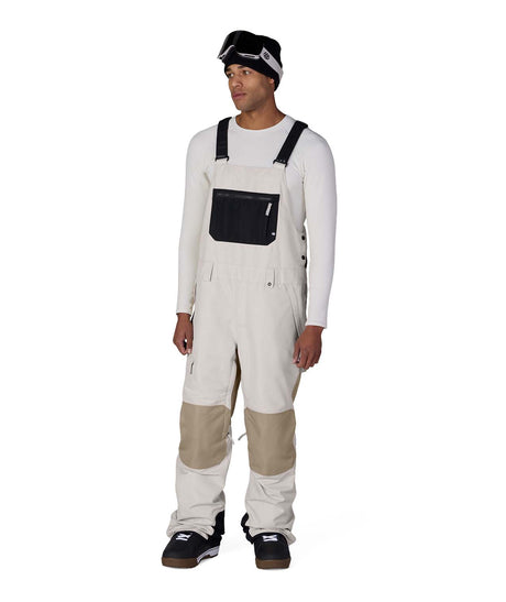 MHotLapInsulatedBib- A man modeling off-white snow bib overalls with beige knee patches, worn over a white long-sleeve shirt and paired with black snow boots and goggles, on a solid white background.