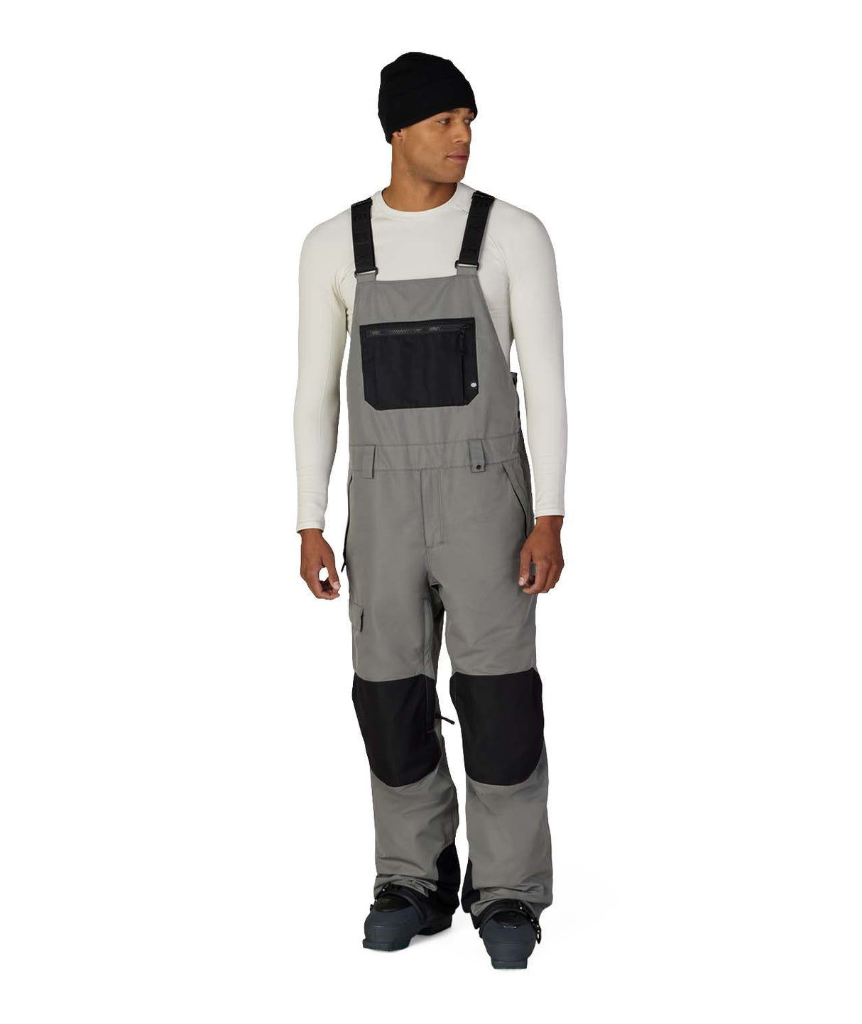MHotLapInsulatedBib- A man modeling gray bib snow pants with black knee patches and a black chest pocket, worn over a white long-sleeve shirt, black beanie, and black boots, on a white studio background.