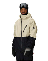 MHydraThermagraphJacket-A man wearing a color-blocked ski jacket in beige and black, along with black snow pants, gloves, and ski goggles, photographed against a solid white background.