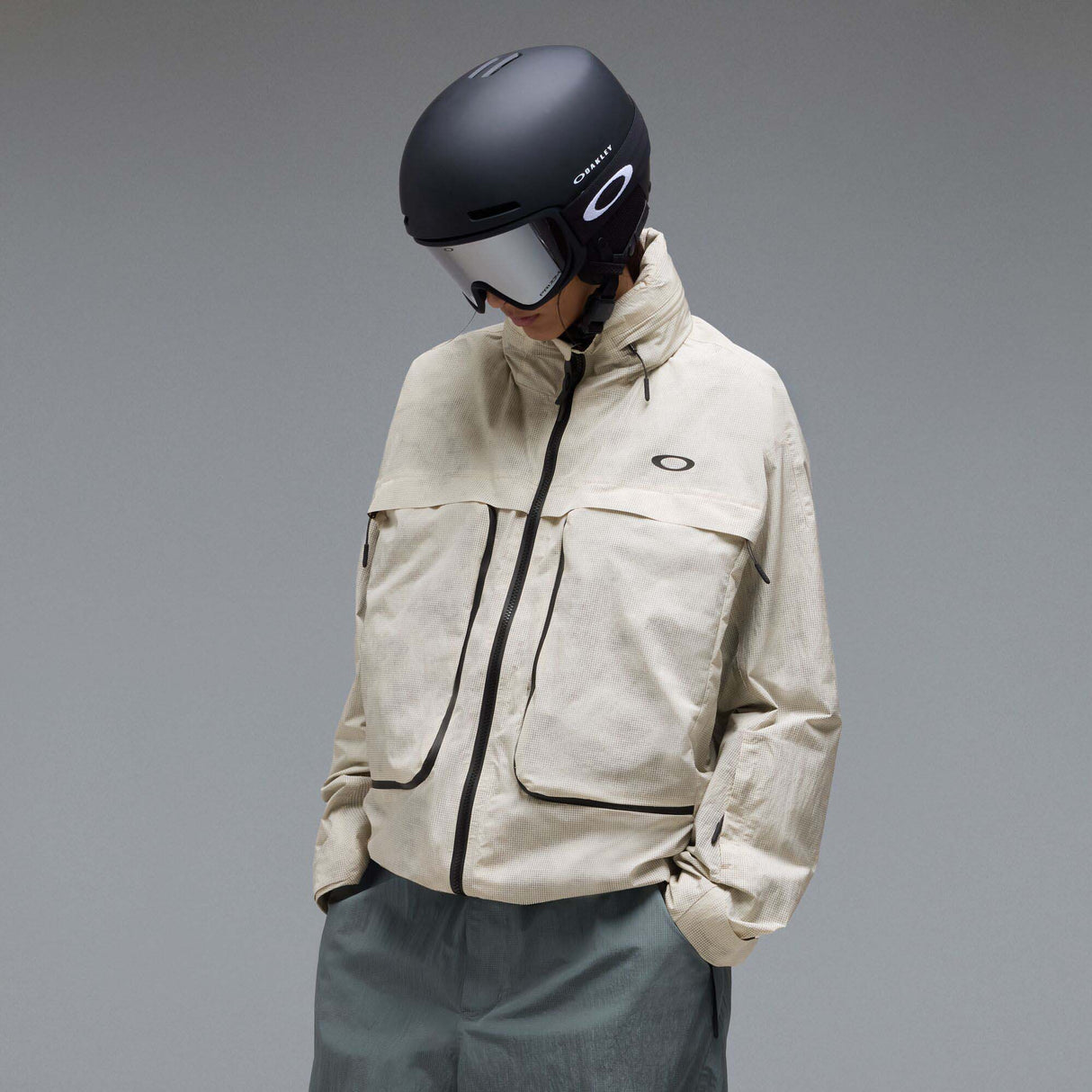 MMthodGlowShellJacket-A light beige technical jacket with black zippers, black piping details, and an Oakley logo on the left chest, displayed against a neutral gray background.