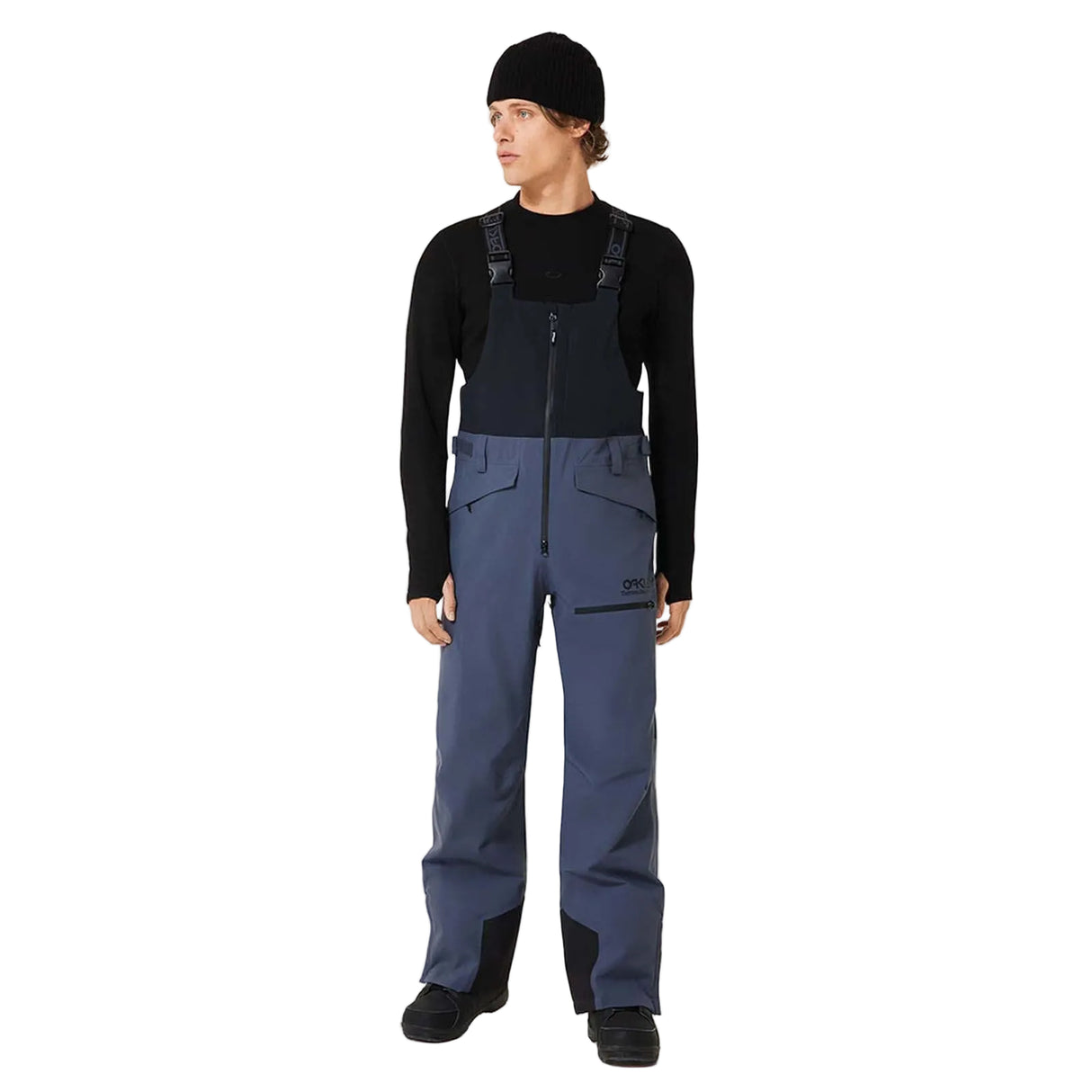 MTNPShellBib-Person wearing a black long-sleeve shirt, black beanie, and blue bib overalls on a white background