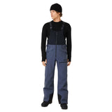 MTNPShellBib-Person wearing a black long-sleeve shirt, black beanie, and blue bib overalls on a white background