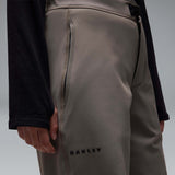 MWintertideSoftshellPant-A pair of gray snow pants with a black belt and a small black Oakley logo on the left thigh, displayed against a neutral gray gradient background.