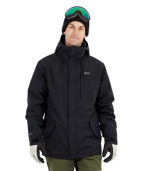 NateJacket-A man wearing a black XTM snowboard jacket, black beanie with green-tinted goggles, black gloves, and olive pants, standing against a solid white background.