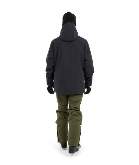 NateJacket-A man wearing a black XTM snowboard jacket, black beanie with green-tinted goggles, black gloves, and olive pants, standing against a solid white background.