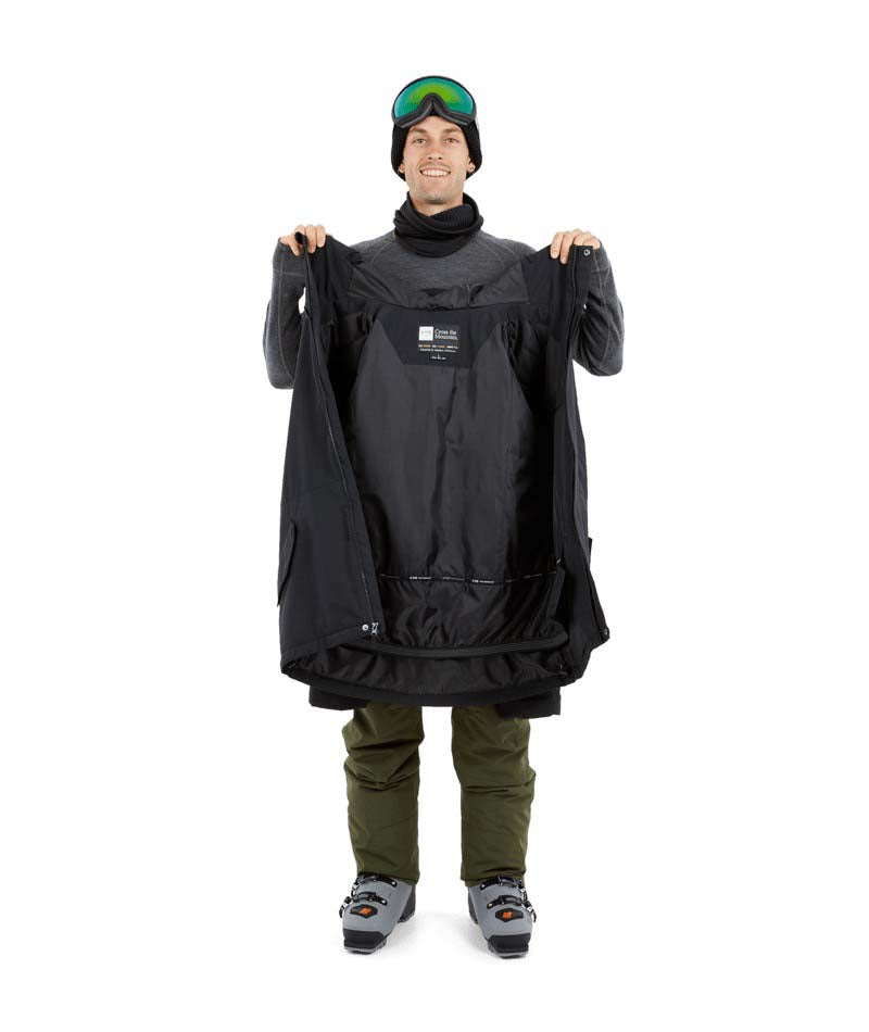 NateJacket-A man wearing a black XTM snowboard jacket, black beanie with green-tinted goggles, black gloves, and olive pants, standing against a solid white background.