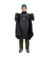 NateJacket-A man wearing a black XTM snowboard jacket, black beanie with green-tinted goggles, black gloves, and olive pants, standing against a solid white background.