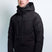 OrionMensInsulatedJacket-A man wearing a black hooded ski jacket with multiple zippered pockets and bright turquoise pants, standing against a light gray studio background.