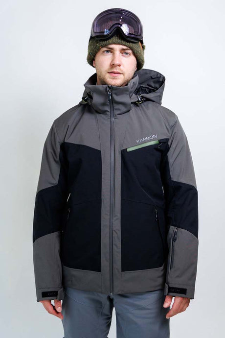 OrionMensInsulatedJacket- A man wearing a Karbon ski jacket in gray and black, a green beanie, and ski goggles, standing against a light gray studio background.