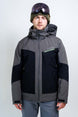 OrionMensInsulatedJacket- A man wearing a Karbon ski jacket in gray and black, a green beanie, and ski goggles, standing against a light gray studio background.