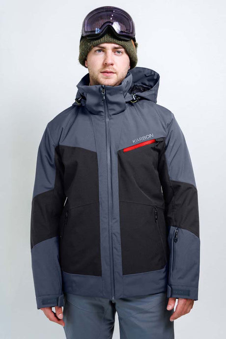 OrionMensInsulatedJacket-A man wearing a Karbon ski jacket in dark gray and black, with a green beanie, ski goggles, and gray pants, photographed against a light gray studio background.